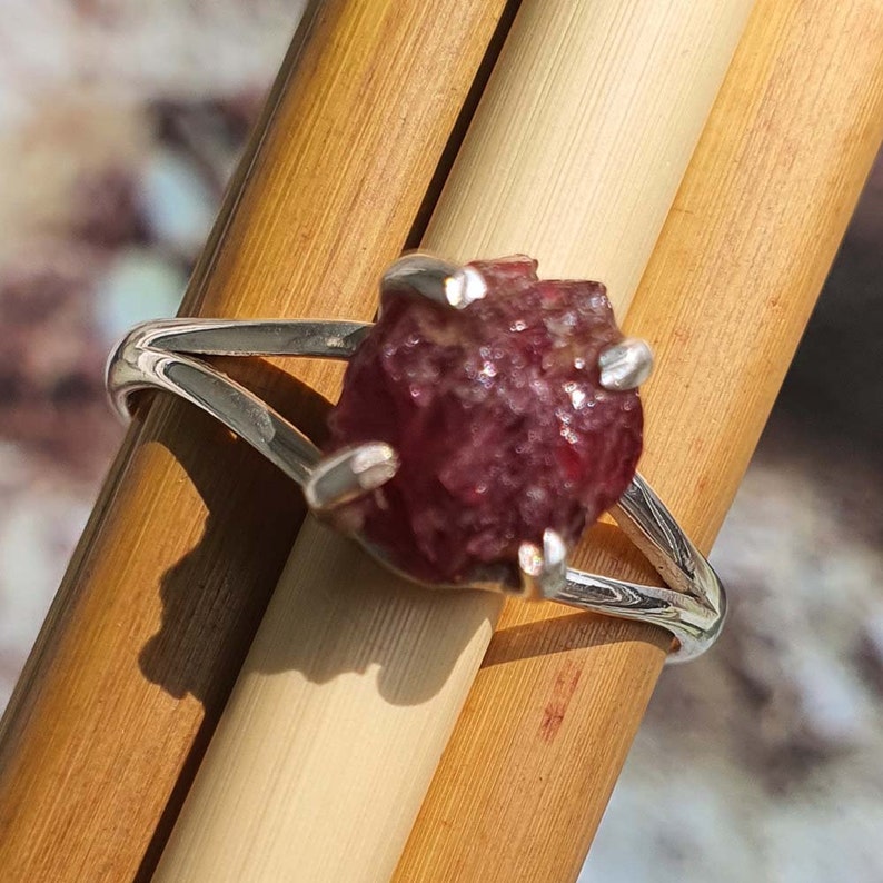 Puede incluir: Un anillo de plata con una piedra preciosa de rub&iacute; rojo en bruto engastada en el centro. El rub&iacute; es de color rojo intenso y tiene una textura rugosa y natural.