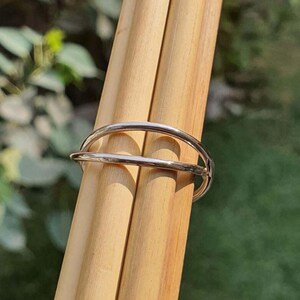 May include: A polished silver-toned metal ring featuring a double-band design. The ring consists of two thin, smooth, parallel bands that curve around the finger, connected at one side to form a single continuous piece of jewelry with a reflective finish.