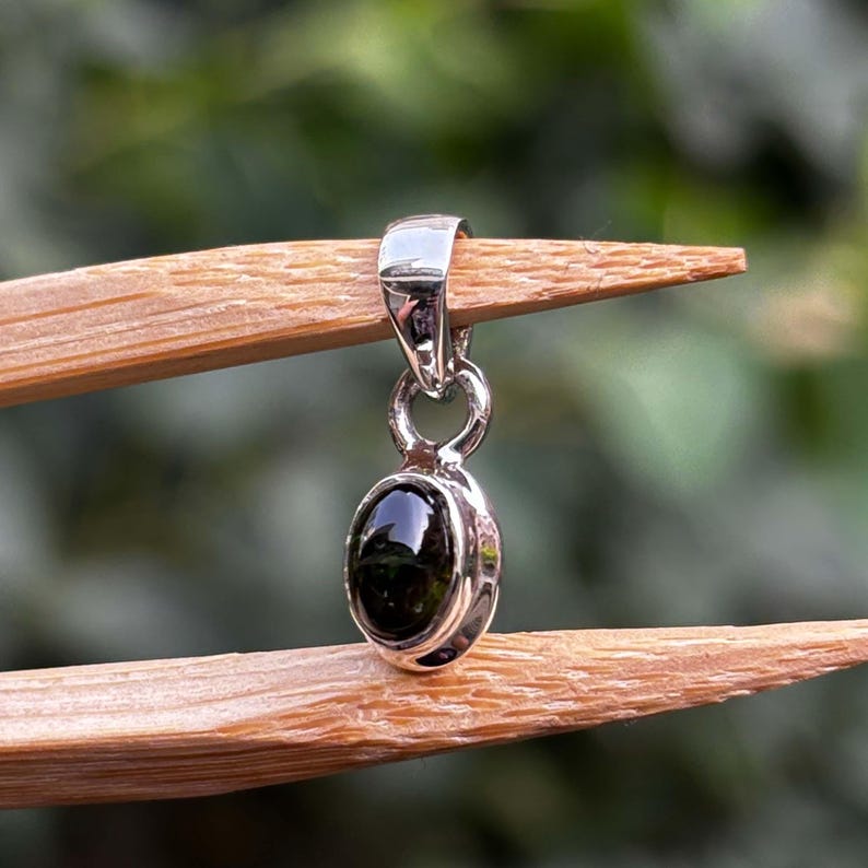May include: A silver pendant featuring a small, polished, oval-shaped black gemstone set in a smooth metal bezel. The pendant includes a simple, rounded bail at the top for attachment to a chain, showcasing a clean and reflective metallic finish.