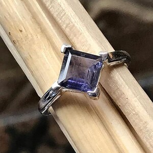 Pode incluir: Um anel de prata com uma pedra preciosa roxa escura de corte quadrado, fixada por quatro garras. A banda &eacute; de prata polida e a pedra apresenta uma superf&iacute;cie facetada que reflete a luz, exibindo um tom violeta escuro com nuances azuis.
