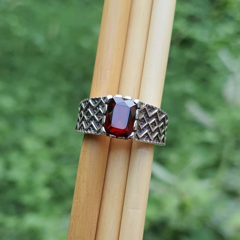 May include: A silver ring with a red gemstone set in a geometric pattern. The ring is on a wooden background.