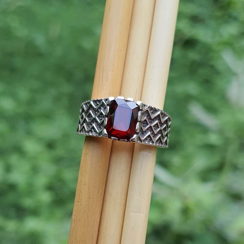 May include: A silver ring with a red gemstone set in a geometric pattern. The ring is on a wooden background.