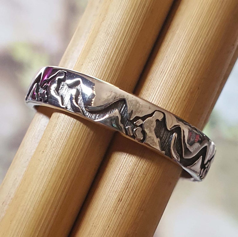 May include: A silver ring with a detailed design of mountain peaks. The ring is on a wooden surface.