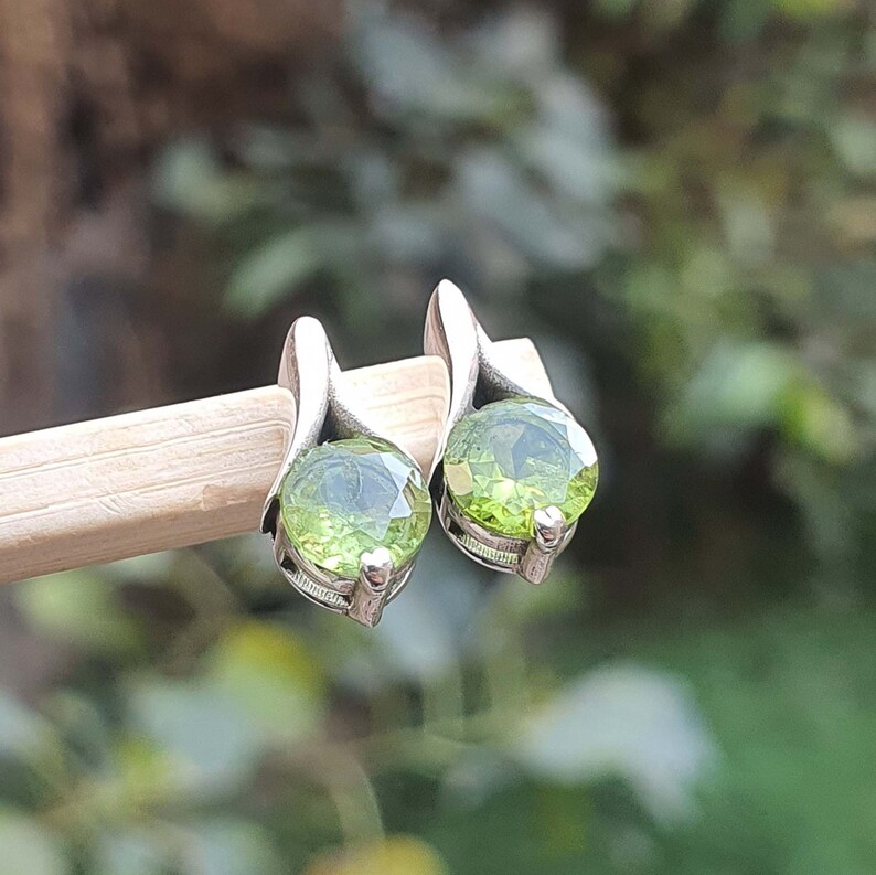 May include: A pair of silver earrings with green gemstone accents. The earrings are shaped like a leaf and have a round, faceted gemstone in the center.