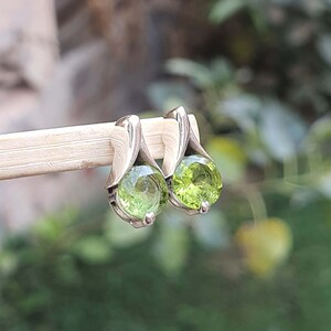May include: A pair of silver earrings with green gemstone accents. The earrings are in the shape of a teardrop with a round gemstone in the center.
