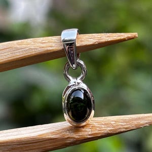 May include: A silver pendant featuring a polished, oval-shaped dark green gemstone set in a smooth metal bezel. The pendant includes a small circular loop connecting the stone setting to a larger, tapered silver bail at the top for hanging.
