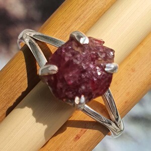 Puede incluir: Un anillo de plata con una gran piedra preciosa roja oscura, en bruto. La piedra est&aacute; engastada en una montura de garras y la banda del anillo es una banda simple y dividida.