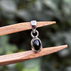May include: A small silver pendant featuring a polished, oval-shaped black gemstone set in a smooth metal bezel. The pendant includes a simple silver bail at the top for attachment to a chain, showcasing a clean, reflective metallic finish.