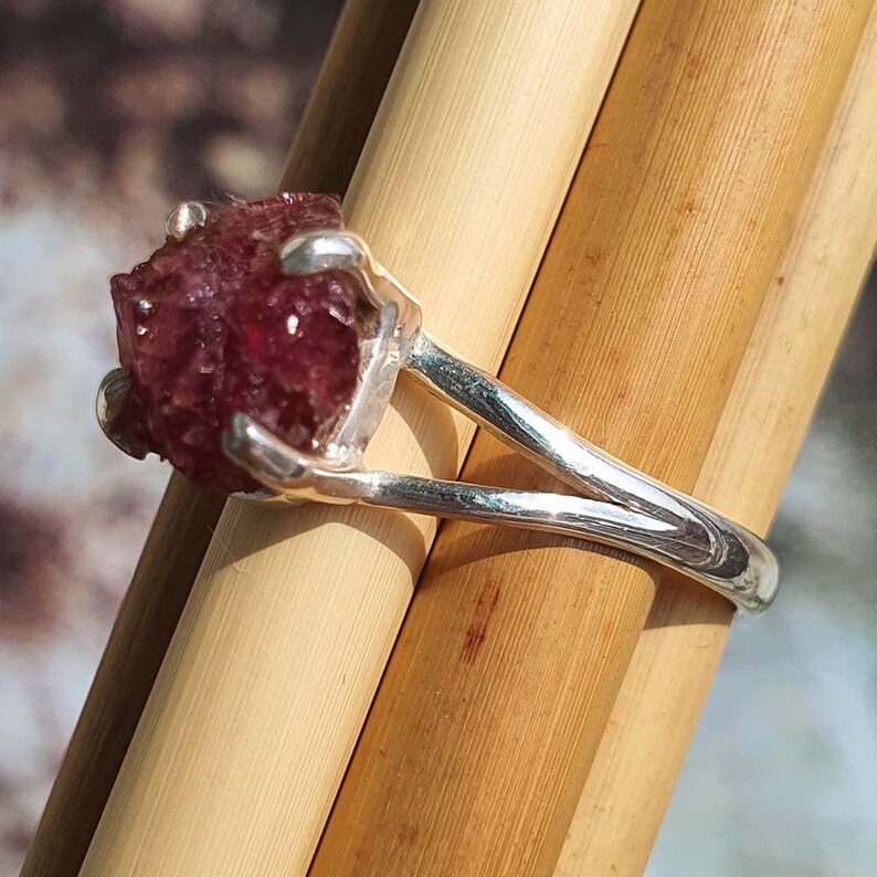 Puede incluir: Un anillo de plata con una piedra preciosa facetada de color rojo oscuro engastada en una montura de garras. El anillo tiene un dise&ntilde;o de ca&ntilde;a dividida.