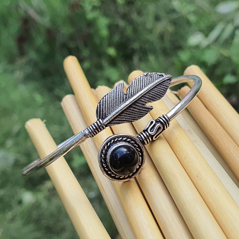 Op de afbeelding: Een zilveren armband met een zwarte steen en een verenontwerp. De armband ligt op een achtergrond van houten stokjes.