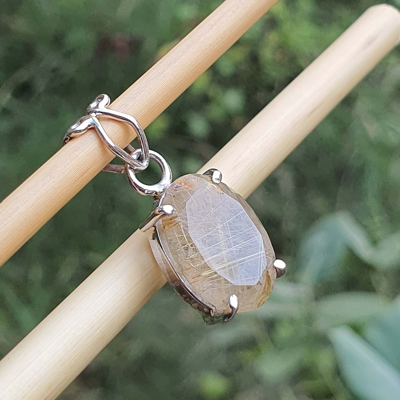 May include: A silver pendant featuring a clear, oval-shaped gemstone with gold inclusions. The pendant has a delicate chain attached to the top.