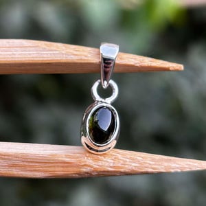 May include: A silver pendant featuring a smooth, oval-shaped dark green gemstone set in a polished metal bezel. The pendant includes a simple silver bail at the top for attachment to a chain, showcasing a clean and reflective metallic finish.