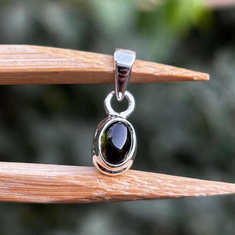 May include: A silver pendant featuring a smooth, oval-shaped dark green gemstone set in a polished metal bezel. The pendant includes a simple silver bail at the top for attachment to a chain, showcasing a clean and reflective metallic finish.
