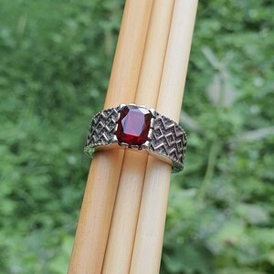 May include: A silver ring with a red gemstone set in a geometric pattern. The ring is on a wooden surface.