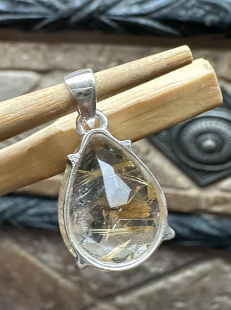 May include: Silver pendant featuring a teardrop-shaped clear quartz crystal with gold inclusions. The pendant is approximately 1 inch in length.