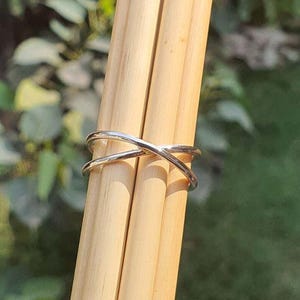 May include: A polished silver-toned ring featuring a crossover design where two thin bands intersect to form an X shape. The ring is positioned horizontally around three light-colored wooden dowels, showcasing its smooth, reflective metallic surface.