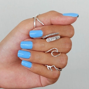 Puede incluir: Una mano con varios anillos de plata. Los anillos tienen un dise&ntilde;o de ola, un dise&ntilde;o de flor y un dise&ntilde;o de hoja. Las u&ntilde;as est&aacute;n pintadas de un color azul claro.