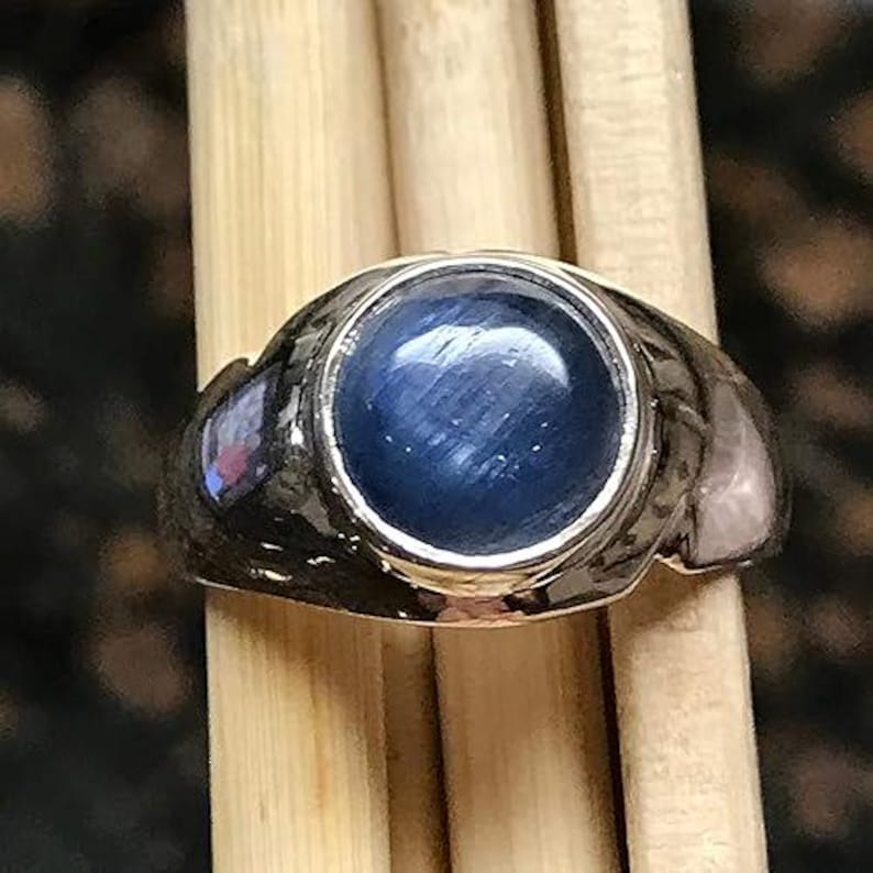 Puede incluir: Un anillo de tono plateado con una piedra preciosa central redonda, tallada en cabuj&oacute;n, de color azul oscuro con un efecto de chatoyance en forma de estrella. La banda de metal tiene un acabado pulido y un dise&ntilde;o ancho y c&oacute;nico.