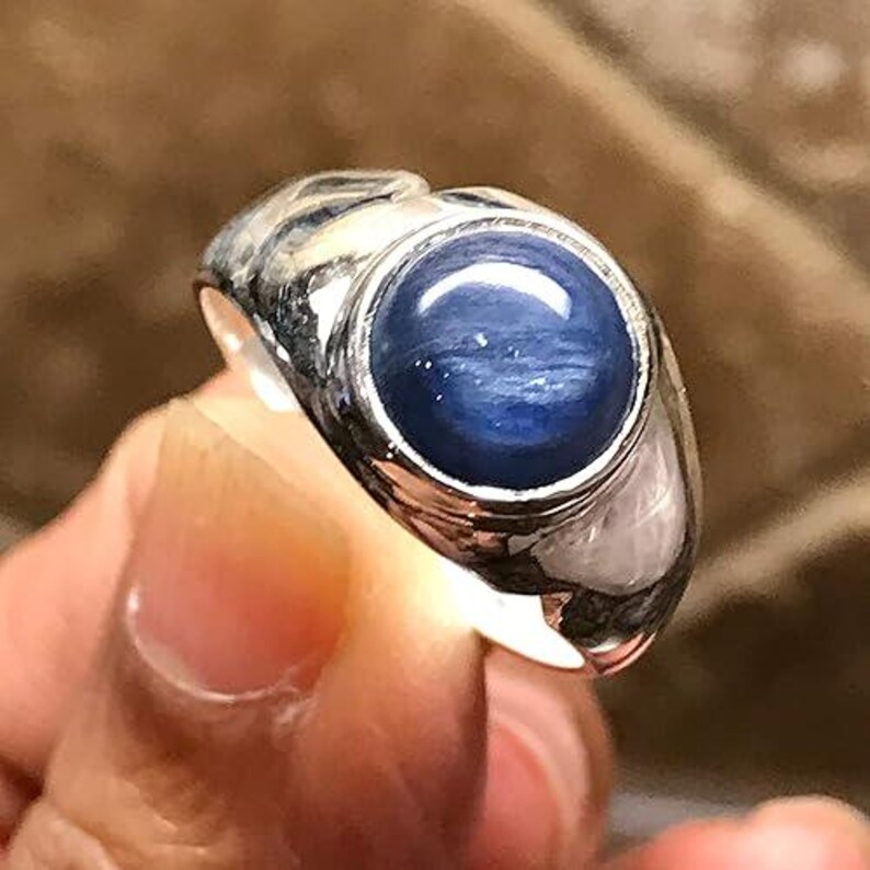Puede incluir: Un anillo de plata con una piedra preciosa cabuj&oacute;n redonda, pulida y de color azul intenso, engastada en un bisel de metal liso. La banda tiene una superficie ancha y reflectante con un dise&ntilde;o curvo y escultural.