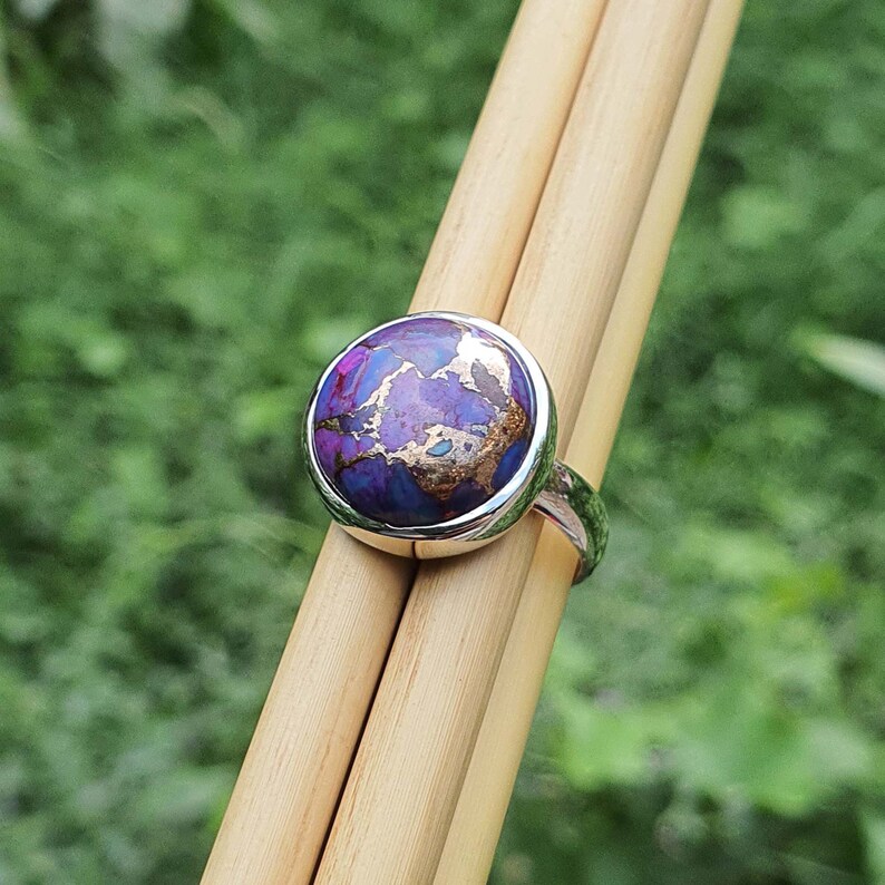 May include: A silver ring with a round purple and gold gemstone. The ring is set on a wooden surface.