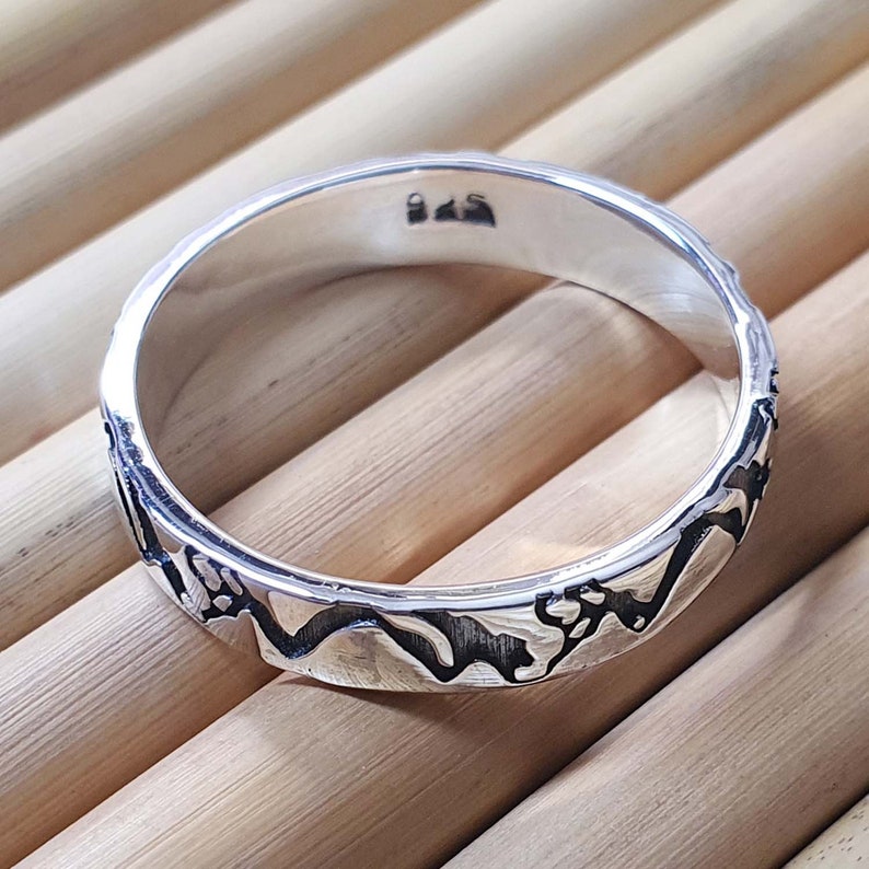 May include: A silver band ring with a textured design of mountain peaks. The inside of the band is stamped with the number "925".
