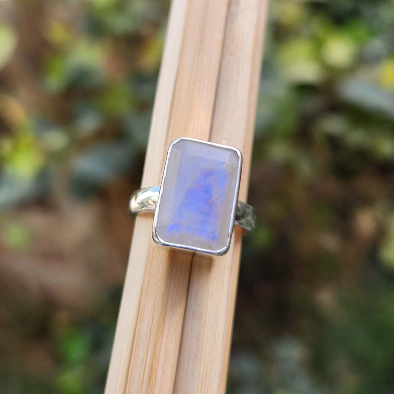 K&ouml;nnte beinhalten: Ein silberner Ring mit einem rechteckigen, schillernden Mondstein. Der Stein hat eine blaue und wei&szlig;e Farbe.