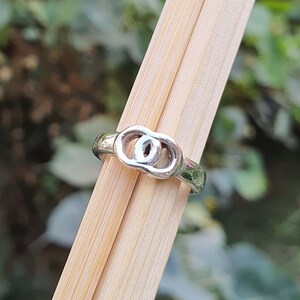 Könnte beinhalten: Ein silberner Ring mit einem Doppeloval-Design. Der Ring ist vor einem Hintergrund aus Holzstäben und grünem Laub platziert, was eine natürliche Ästhetik erzeugt. Die polierte Oberfläche des Rings reflektiert das Licht.