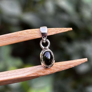 May include: A small silver pendant featuring a polished, oval-shaped black gemstone set in a smooth metal bezel. The pendant includes a simple silver bail at the top for attachment to a chain, showcasing a clean, reflective metallic finish.