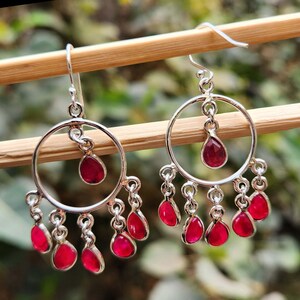 May include: Silver hoop earrings with dangling red gemstone drops. The earrings have a circular design with a single larger red gemstone drop in the center of each hoop. The earrings are accented with smaller red gemstone drops hanging from the bottom of each hoop.