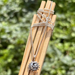 K&ouml;nnte beinhalten: Eine silberfarbene Metallkette mit einem Anh&auml;nger, der ein kombiniertes Sonnen- und Mondgesicht zeigt. Der Anh&auml;nger h&auml;ngt an einer feinen Gliederkette, die &uuml;ber eine helle Holzstruktur drapiert ist. Das Metall hat eine polierte Oberfl&auml;che.