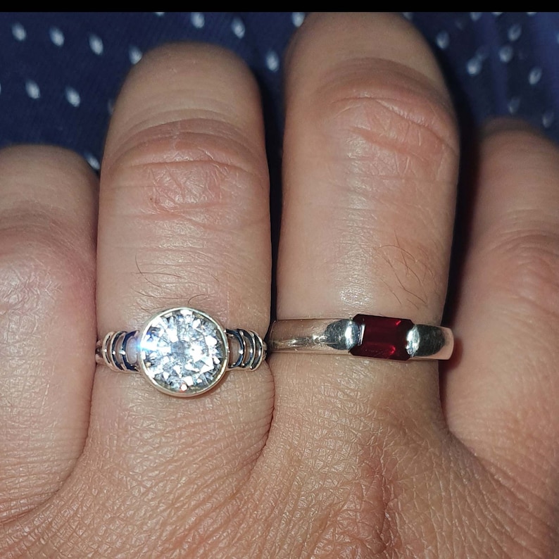 May include: Two silver rings with gemstones. The ring on the left has a large, clear, round gemstone. The ring on the right has a smaller, rectangular, red gemstone.