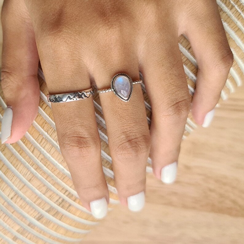 May include: Two silver rings on a finger. The top ring features a teardrop-shaped moonstone. The bottom ring has a textured design.