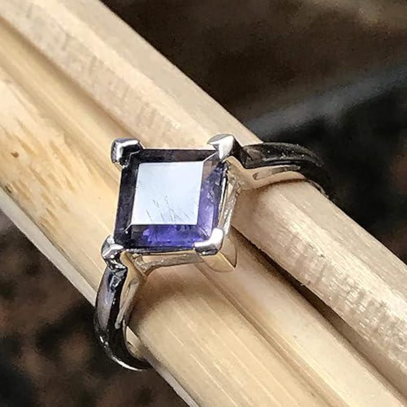 Pode incluir: Um anel de prata com uma pedra preciosa roxa escura de corte quadrado, montada em quatro garras. A banda &eacute; de prata polida e a pedra apresenta uma superf&iacute;cie facetada com reflexos de luz vis&iacute;veis na sua face violeta escura e transl&uacute;cida.