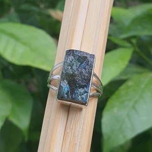 Puede incluir: Un anillo de plata con una piedra rectangular de labradorita verde oscuro. La piedra tiene una calidad brillante e iridiscente.
