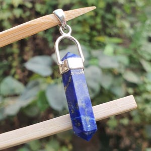 Pode incluir: Um pingente de prata com uma pedra preciosa de lápis-lazúli azul em forma pontiaguda. O pingente está suspenso de uma fivela de prata.