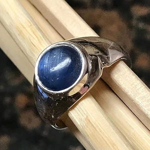 Puede incluir: Un anillo de tono plateado con una piedra preciosa cabuj&oacute;n redonda de color azul profundo, engastada en una banda de metal pulido. El anillo se muestra sobre una superficie de madera clara, resaltando el brillo del metal y la piedra.