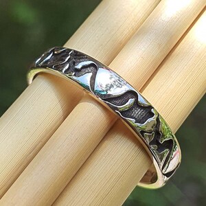 May include: A silver band ring with a textured design of mountains and trees. The ring is shown on a wooden surface.