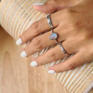 May include: A hand wearing three silver rings. The middle ring features a teardrop-shaped moonstone.