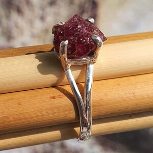 Puede incluir: Un anillo de plata con una piedra preciosa facetada de color rojo oscuro engastada en garras. El anillo tiene un dise&ntilde;o &uacute;nico con una banda dividida.