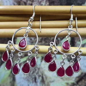 May include: A pair of silver hoop earrings with dangling red gemstone drops. The earrings have a circular design with a single red gemstone drop in the center of each hoop. The earrings are adorned with multiple smaller red gemstone drops hanging from the bottom of each hoop.