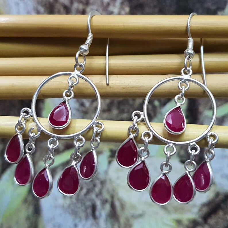 May include: A pair of silver hoop earrings with dangling red gemstone drops. The earrings have a circular design with a single red gemstone drop in the center of each hoop. The earrings are adorned with multiple smaller red gemstone drops hanging from the bottom of each hoop.