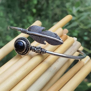 Op de afbeelding: Zilveren armband met een zwarte steen en een veerbedel. De armband is open en verstelbaar.
