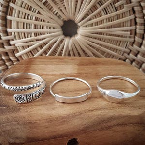 Puede incluir: Tres anillos de plata sobre una superficie de madera. Los anillos son de diferentes estilos. Un anillo es una banda simple, otro es una banda con un dise&ntilde;o decorativo y el tercero es un anillo con una forma ovalada redondeada.