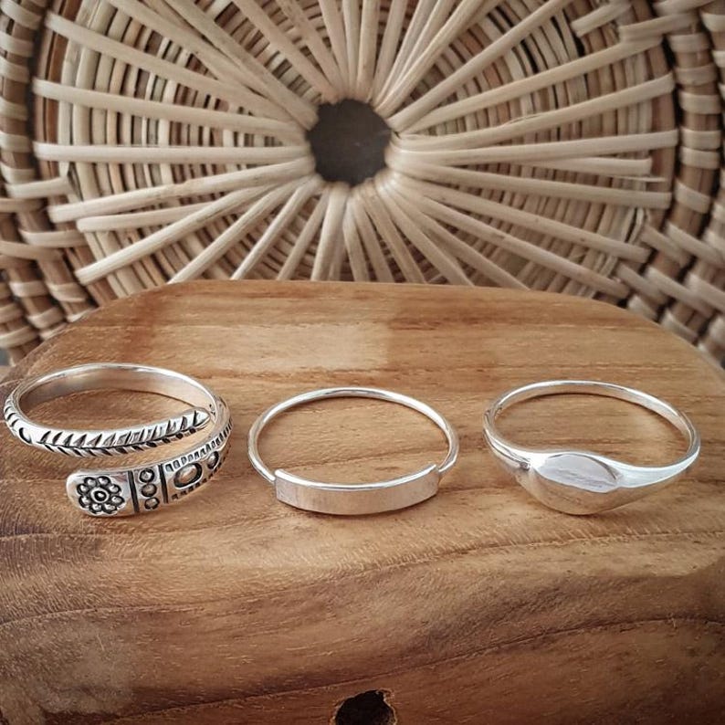 Puede incluir: Tres anillos de plata sobre una superficie de madera. Los anillos son de diferentes estilos. Un anillo es una banda simple, otro es una banda con un dise&ntilde;o decorativo y el tercero es un anillo con una forma ovalada redondeada.