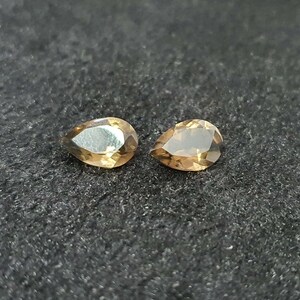 May include: Two pear-shaped gemstones, one with a light brown color and the other with a darker brown color, are displayed on a black surface.