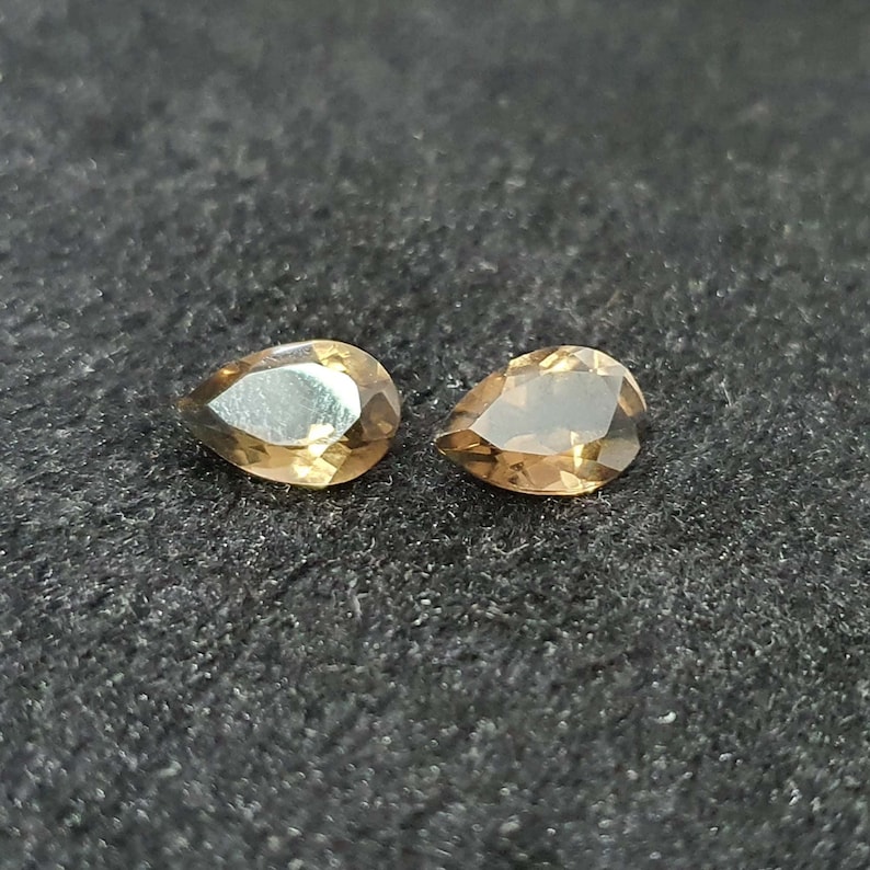 May include: Two pear-shaped gemstones, one with a light brown color and the other with a darker brown color, are displayed on a black surface.