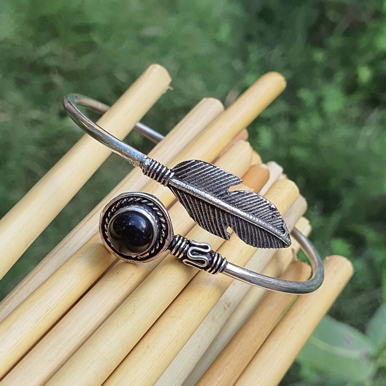 Op de afbeelding: Een zilveren armband met een zwarte edelsteen en een veerbedel. De armband is open en heeft een eenvoudig ontwerp.