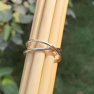 May include: A polished silver-toned ring featuring a crossover design with two thin, smooth bands that intersect at the center. The ring is positioned around a bundle of light-colored, cylindrical wooden dowels, showcasing its metallic finish.