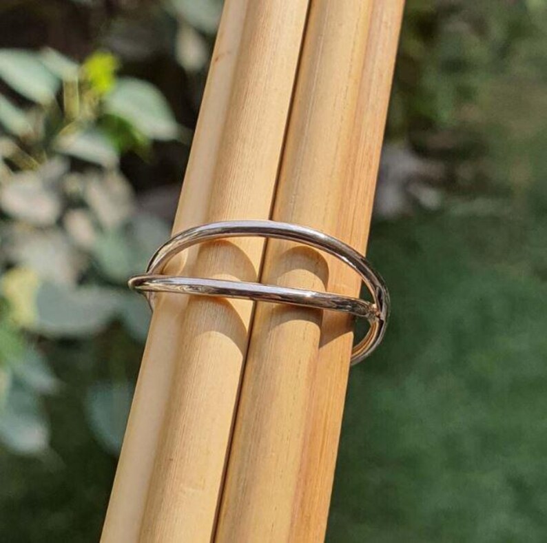 May include: A polished silver-toned metal ring featuring a double-band design. The ring consists of two thin, smooth, parallel bands that curve around the finger, connected at one side to form a single continuous piece of jewelry with a reflective finish.