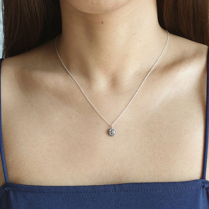 K&ouml;nnte beinhalten: Eine silberfarbene Halskette mit einem kleinen, runden Anh&auml;nger, der ein Sonnen- und Mondgesicht zeigt. Der Anh&auml;nger h&auml;ngt an einer zarten, d&uuml;nnen silberfarbenen Kette, die auf dem Schl&uuml;sselbein aufliegt. Das Metall hat eine polierte Oberfl&auml;che.